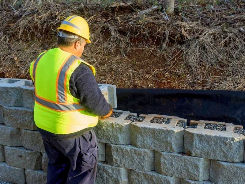 Masonry contractor installing a stone retaining wall with block courses and drainage preparation on a sloped landscape