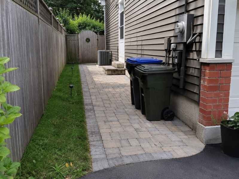 Stone pavers side entrances Madison Alabama - Madison Brick & Stone Paved side yard with black trash and blue-lidded recycling bins beside a house, along a wooden fence toward a gate at the end.