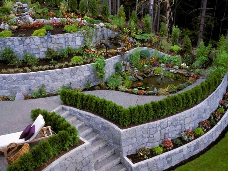 Terraced stone garden with a winding pathway, small pond, and lush shrubs, featuring stone walls and a seating area at the lower left.
